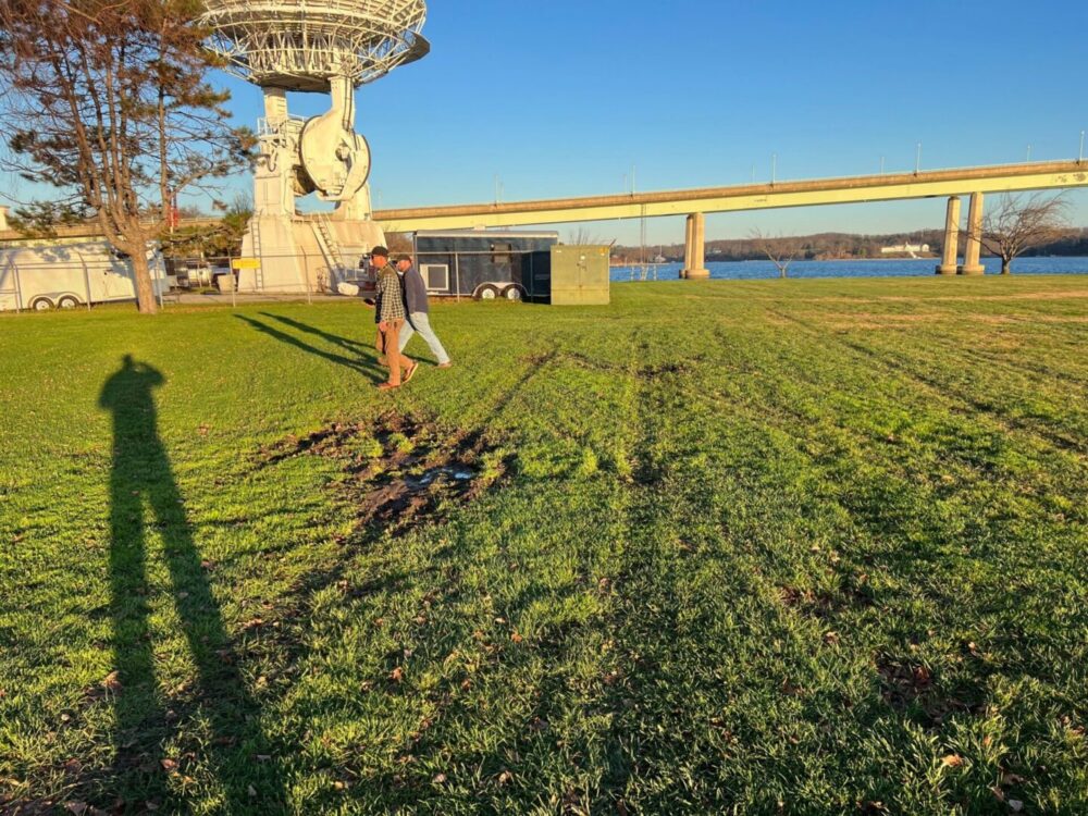 Person working with a rake on a grassy field near a bridge and satellite dish.